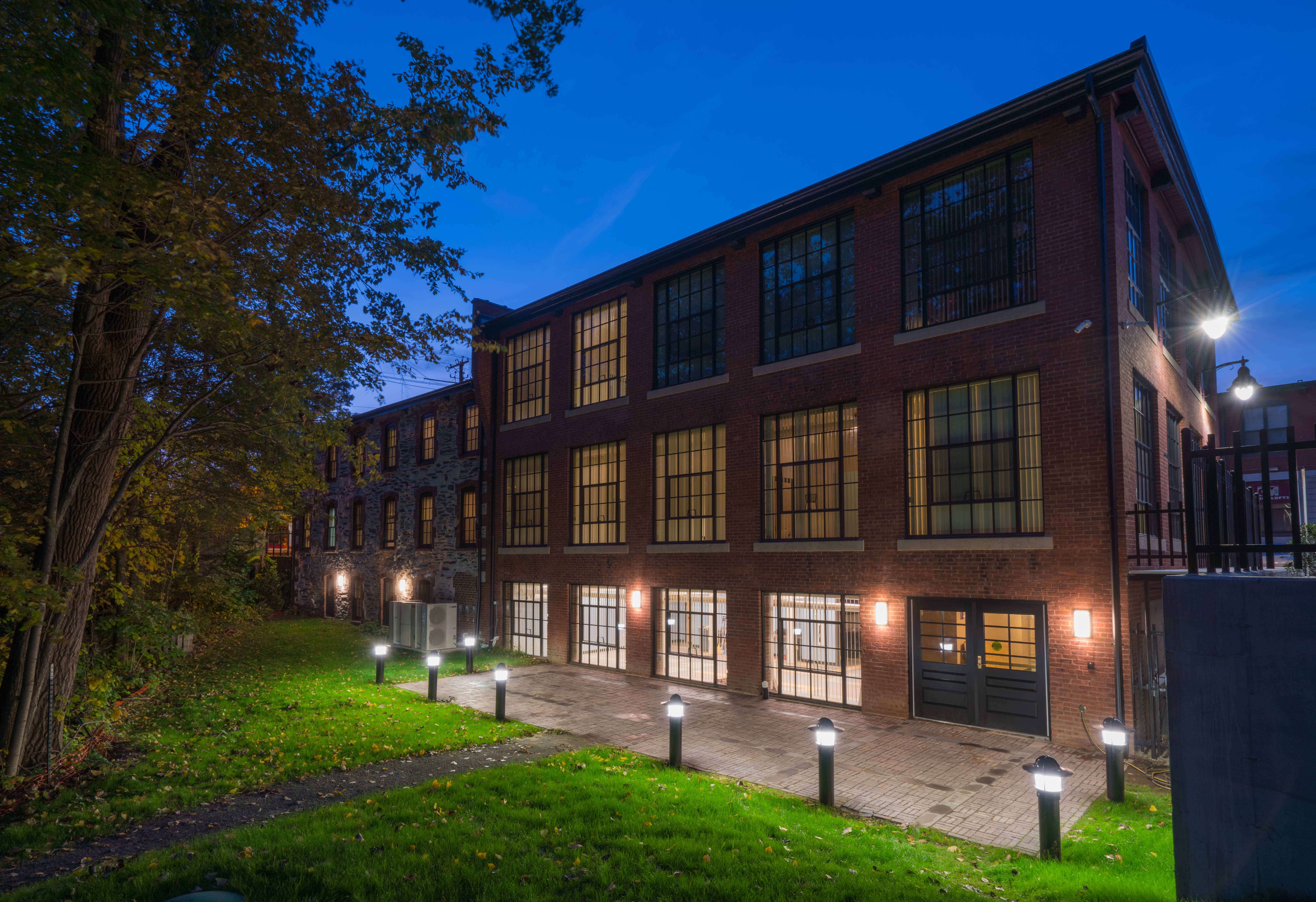 Exterior view of a multi-story brick building with large windows and two doors, walkway lights and green grass surrounding the building.