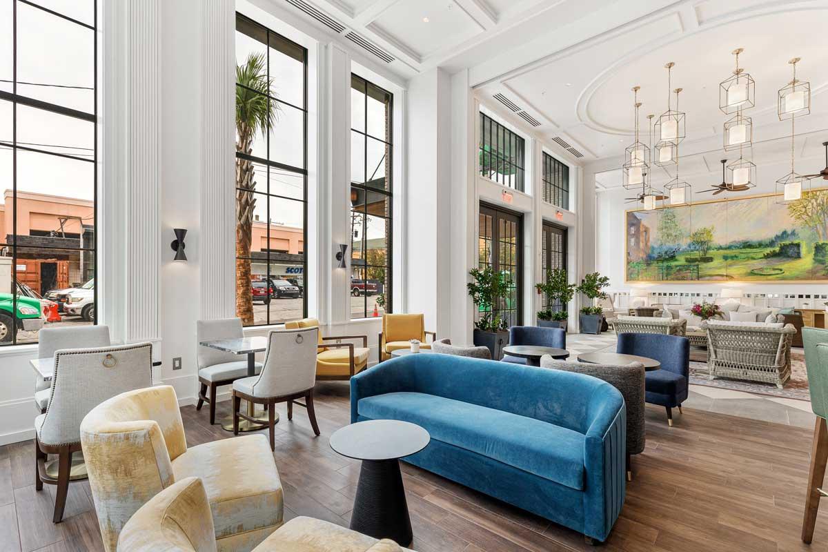 Looking through the floor to ceiling windows from the inside of the Loutrel Hotel, this lobby contains various comfortable individual chairs, as well as couches. Palm trees and cars are visible just outside the windows.