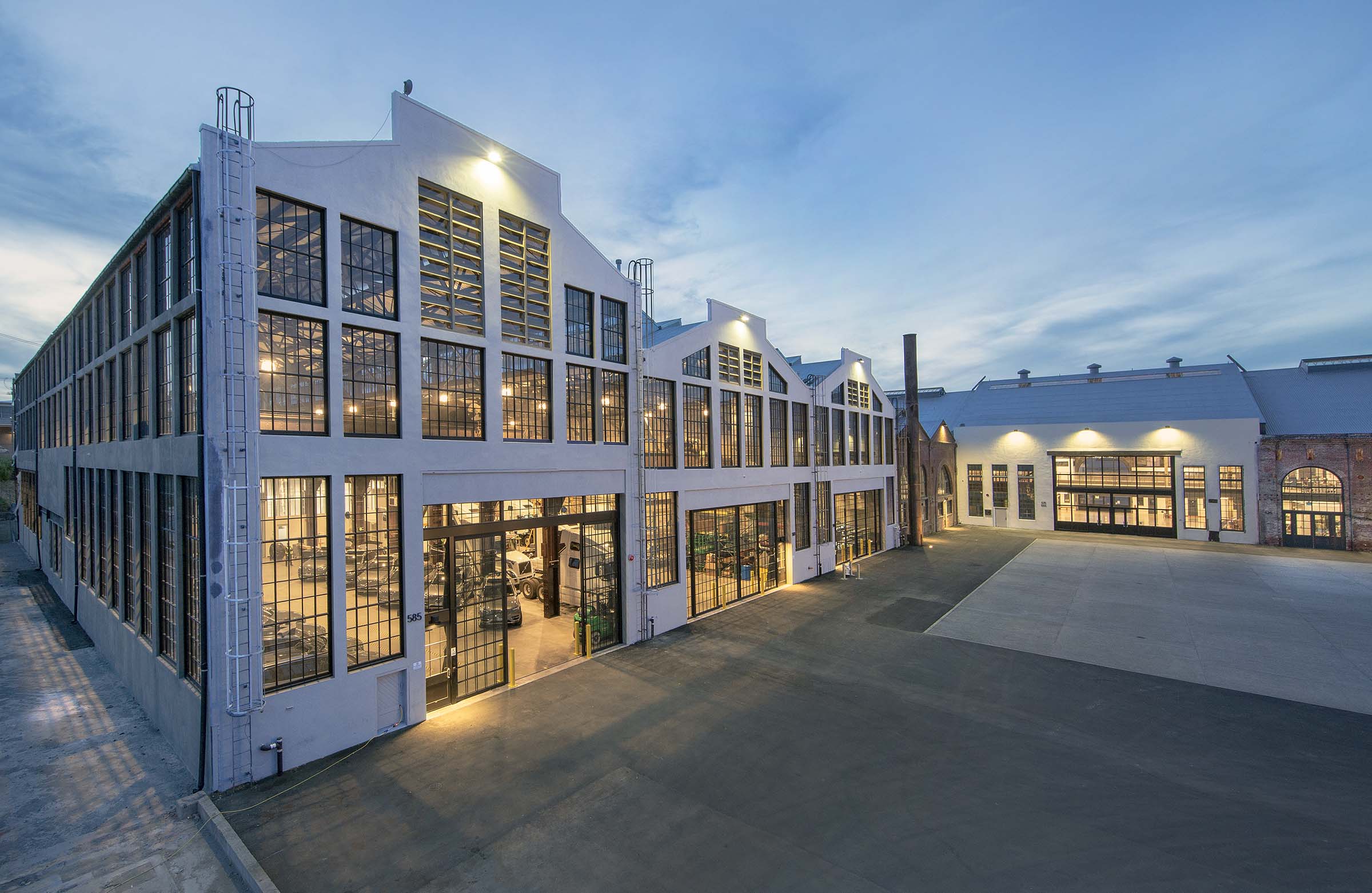 Large commercial building photographed at twilight with floor-to-ceiling windows on multiple sides and white siding, and there is a darkening blue sky in the background.