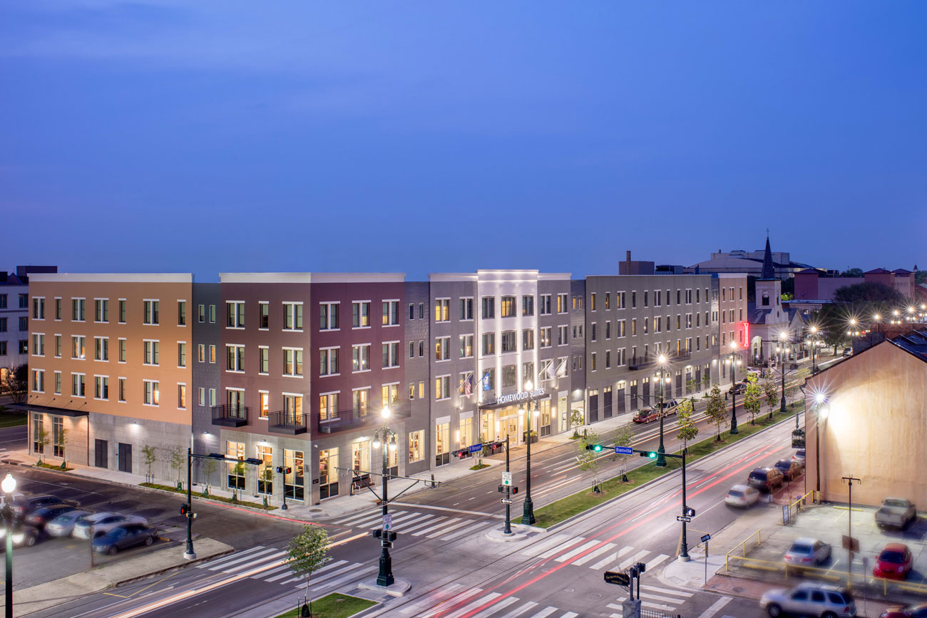 A hotel at night with multiple windows, several filled with light, next to an intersection and multiple street lights.
