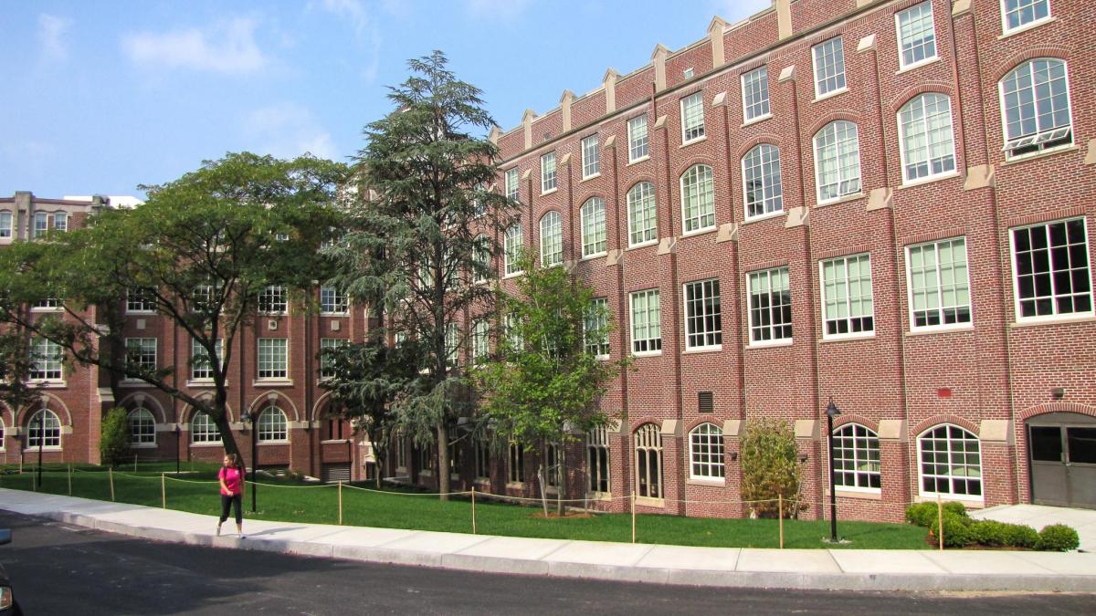 Harkins Hall has a brick exterior with various windows. There is a person wearing black pants and a pink shirt is walking in front of the building.