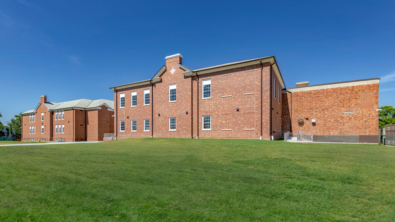 Exterior brick building featuring historic replica windows with a grassy field in front and blue sky in the background.