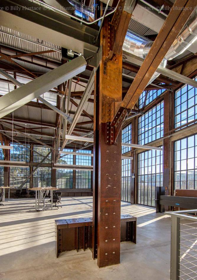 A large indoor room featuring a prominent metal beam, with natural daylight illuminating the space. The floor is visible beneath the beam, showcasing materials like steel and aluminum in the structure.