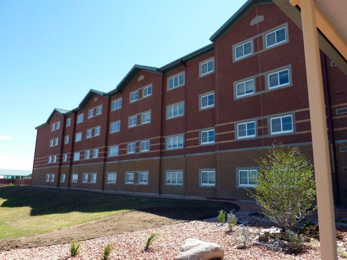 A large building featuring numerous sliding windows, surrounded by greenery including trees and grass, under a clear blue sky.