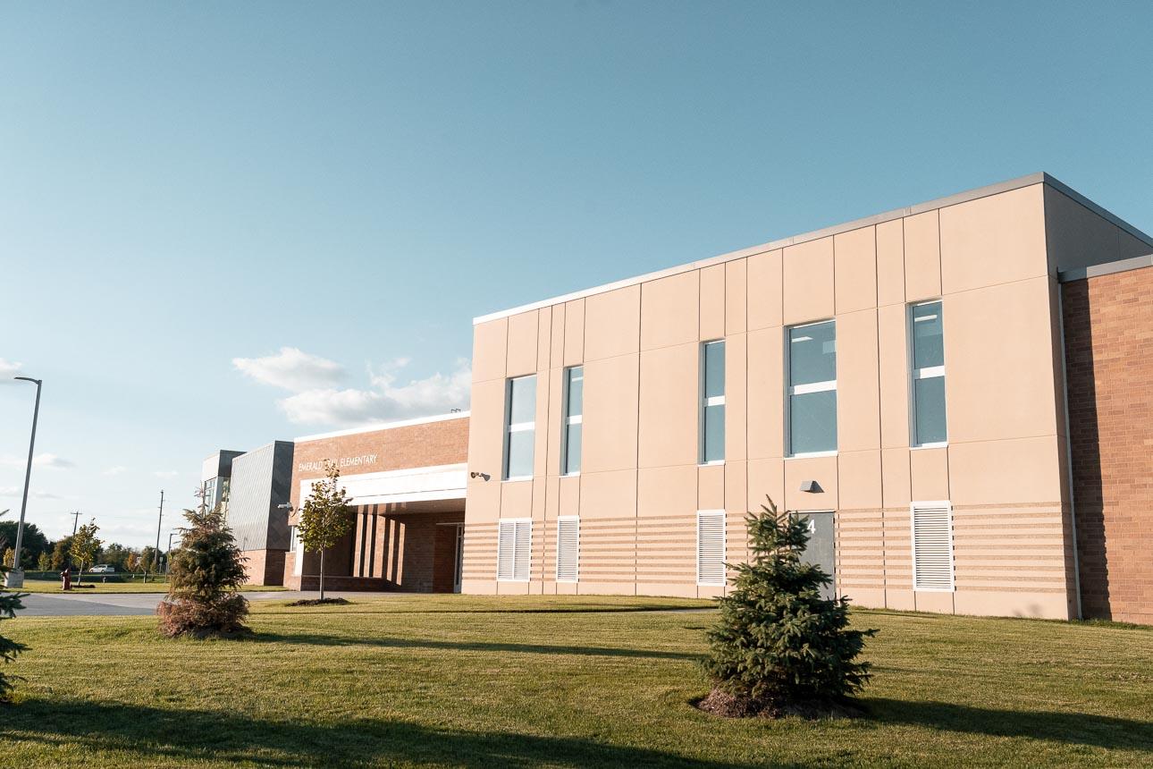 Modern school building with beige and brick facade under a clear blue sky. Manicured lawn and young trees create a peaceful, sunny atmosphere.