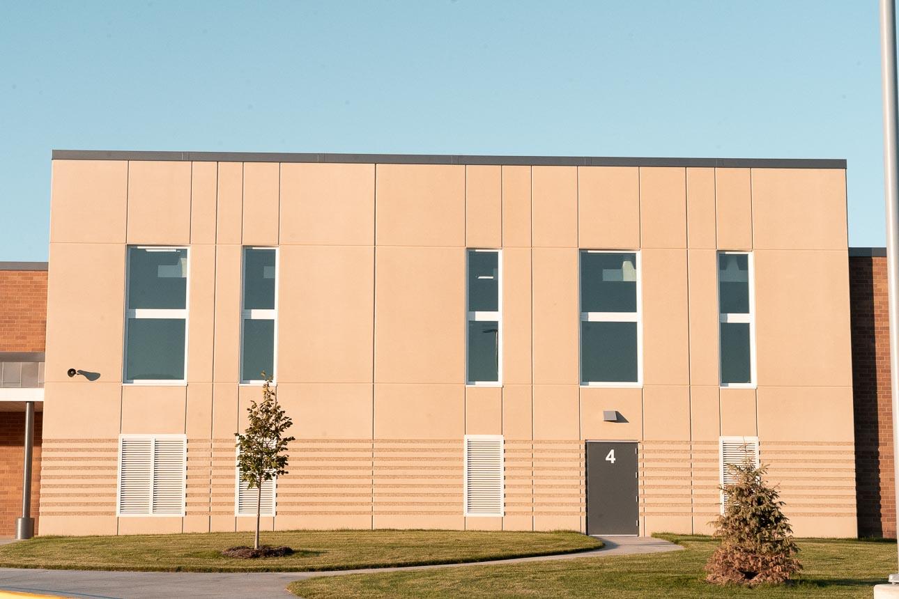 A beige building facade with vertical windows and a door marked "4." It is surrounded by a neatly trimmed lawn and two young trees under a clear sky.